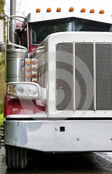 Classic wet big rig red semi truck with chrome grille and bumper standing on the street in raining weather