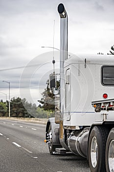 Classic white powerful American bonnet big rig semi truck with tall vertical chrome pipe and flat bed semi trailer running on the