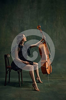 Classical music live performance. Elegant young woman, cellist, passionate musician sitting with cello against dark