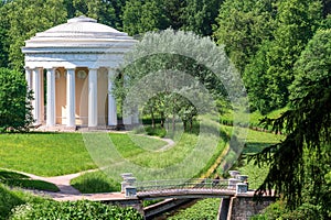 A classical-style temple in a beautiful summer park Green field with white and yellow dandelions outdoors in nature in summer.