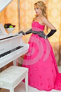 Classy blonde in long red dress is standing next to the piano