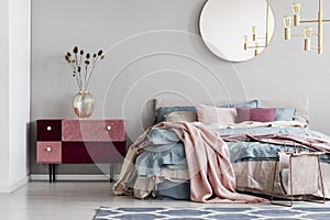 Classy round mirror on grey wall in stylish bedroom interior with warm bed with blue, pastel pink and beige bedding and velvet