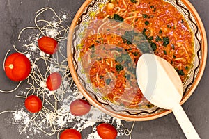 Clay bowl with traditional dish consist of tomatoes with grain noodles on slate and wooden spoon and ingredients near