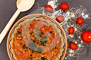 Clay bowl with traditional dish consist of tomatoes with grain noodles on slate and wooden spoon and ingredients near