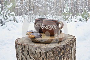 Clay cups with coffee and cookies on a wooden stump in a snowy winter forest