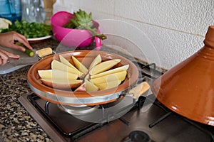 A clay dish on the stove, with slices potato on the kitchen counter. Woman cooking Moroccan tagine
