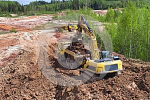 Clay mining. The yellow excavator digs red clay with a large bucket and puts it into the truck