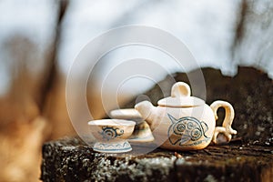 Clay white teapot with blue pattern and two drinking bowls in the forest on the stump. Handwork