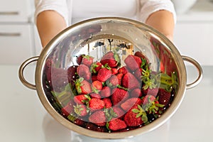 Clean red ripe strawberries in a stainless steel colander are rinsed under water in a woman hand.