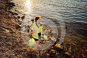 Clean up of pebble wild beach. Top view of young Caucasian woman wearing vest and rubber gloves holds plastic bag with