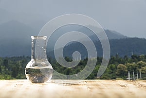 Clean water in a glass laboratory flask on wooden table on mountain background. Ecological concept, the test of purity