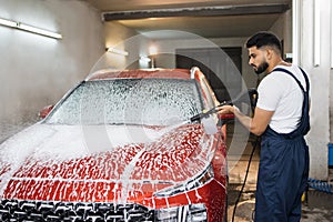 Handsome young bearded man worker, wearing protective clothes and gloves, washing modern red car