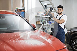 Handsome man worker washing modern red car hood under high pressure water in car wash service