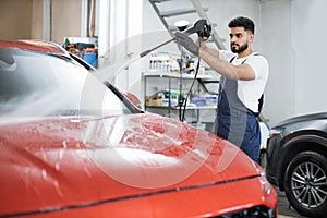 Handsome man worker washing modern red car hood under high pressure water in car wash service