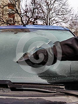 Cleaning car windows in winter. Man scrapes hoarfrost with a plastic scraper from the windshield of a blue car.