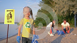 Cleaning coast, portrait of little happy volunteer girl in rubber gloves with trash bag near pointer sign on background