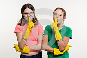 Cleaning, domestic duties and teamwork concept. Two teenage sisters wearing yellow protective gloves show sign quieter, secret