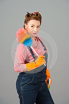 Cleaning lady with colored duster and orange latex gloves in pin-up style. Brunette curl retro styled housekeeper on grey studio