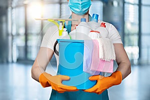 A cleaning lady in a mask holds a bucket of cleaning products