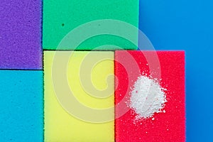 Cleaning powder on a red dishwashing sponge next to multi-colored sponges on a blue background. The concept of homework