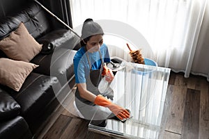 Cleaning service worker in wearing rubber gloves wiping the dust off glass table in living room