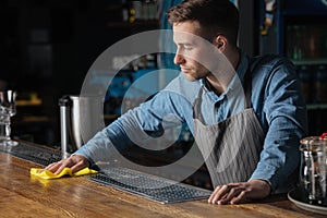 Cleanliness in pub. Professional bartender standing at counter, wiping wooden surface with rag