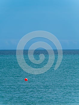 Clear blue sky over a blue sea. An orange buoy on the surface of the water. The horizon is above the water. Minimalistic landscape