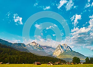 A clear blue-white sky with sun and clouds over the massif of the Bavarian Alps in front of a mountain meadow with a forest and