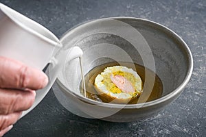 Clear broth is poured around a pate slice of three kinds of fish in a gray ceramic soup bowl, preparation of a gourmet menu,