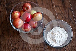 Clear glass bowl of apples and clear glass bowl of flour positioned on a dark wood background