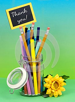 A clear hermetic jar filled with colorful makers and pencils on a graduated blue green blackground with yellow silk flower