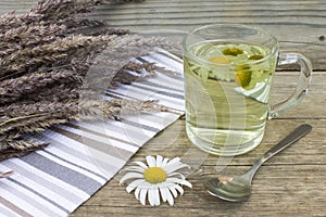 Clear transparent cup of camomile tea on vintage wooden background with dried herbs and daisy flowers.