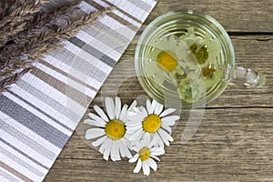 Clear transparent cup of camomile tea on vintage wooden background with dried herbs and daisy flowers.