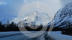 Cleared rural road with snowplow markings, bare trees and snow-covered mountains in background between Fagernes and Brevikeidet. photo