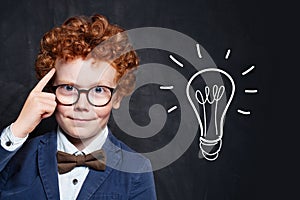 Clever boy in glasses with idea lightbulb on blackboard background photo