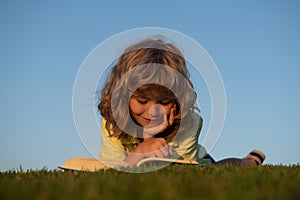 Clever child boy reading book laying on grass on grass and sky background with copy space.