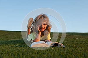 Clever child boy reading book laying on grass on grass and sky background with copy space. Closeup portrait of of a boy