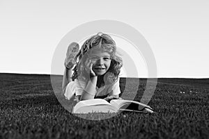 Clever child boy reading book laying on grass on grass and sky background with copy space. Closeup portrait of of a boy