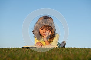 Clever child boy reading book laying on grass on grass and sky background with copy space. Kids imagination, innovation