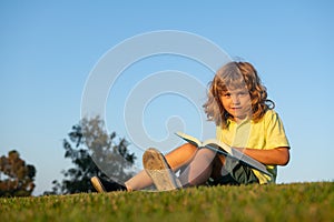 Clever child boy reading book laying on grass and sky background with copy space.