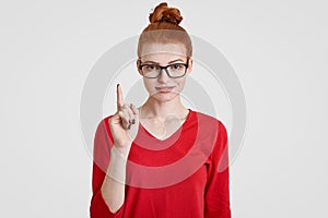 Clever freckled foxy student gets brilliant idea in mind, raises index finger in eureka gesture, wears transparent glasses, red sw