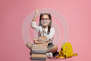 Clever pupil. Smiling girl in glasses with stack of books and backpack knows answer and raises hand up