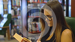 Clever young woman sits on armchair and reads book at the library