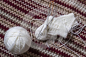 A clew of white wool yarn, knitting needles and unfinished mittens lie on a homemade multi-colored bedspread