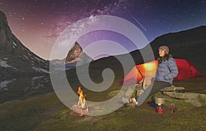 Climber resting next to the tent and fire, while preparing a hot tea, Matterhorn peak behind and the Milky Way