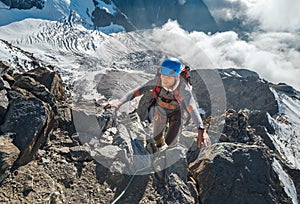 Climber in a safety helmet, harness with backpack ascending a rock wall with Bionnassay Glacier on background and looking at the