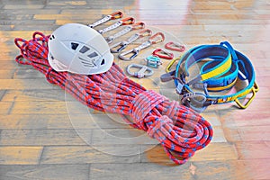 Climbing equipment on wooden background: red dynamic rope with blue stripes, helmet, blue/yellow harness, quickdraws, belay device