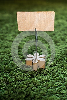 Clip card holder with wooden flower on green grass carpet holding a natural wooden announcement display with empty space for text photo