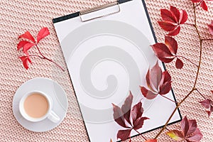 Clipboard, autumn leaves and cup of coffee with milk on pastel pink knitted plaid background. Autumn cozy. Flat lay, top view,