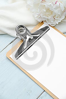 Clipboard with blank paper mockup and peonies on wooden table, top view. Wedding planning concept